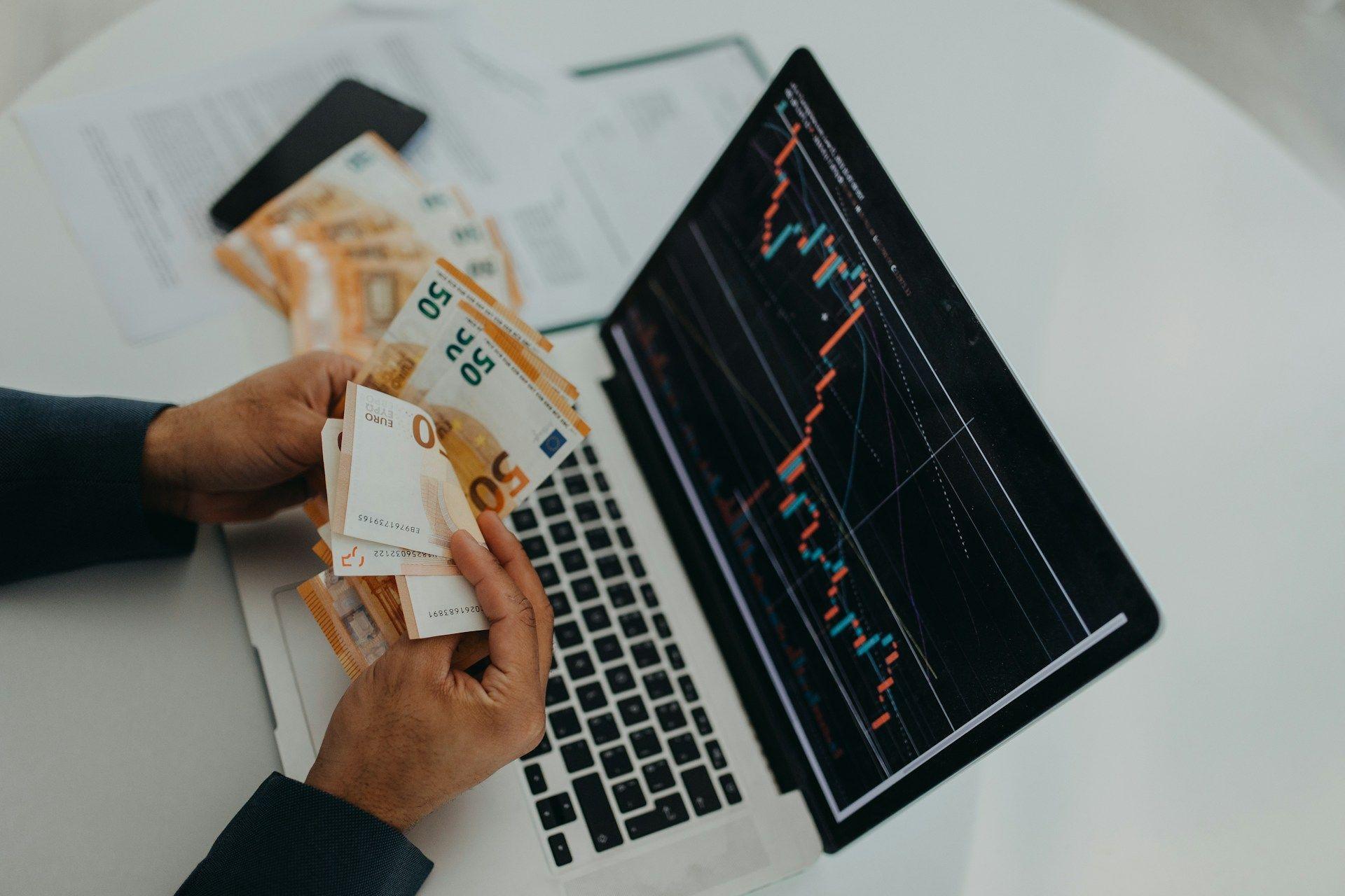 Hands counting Euro banknotes next to a laptop displaying a financial market chart.