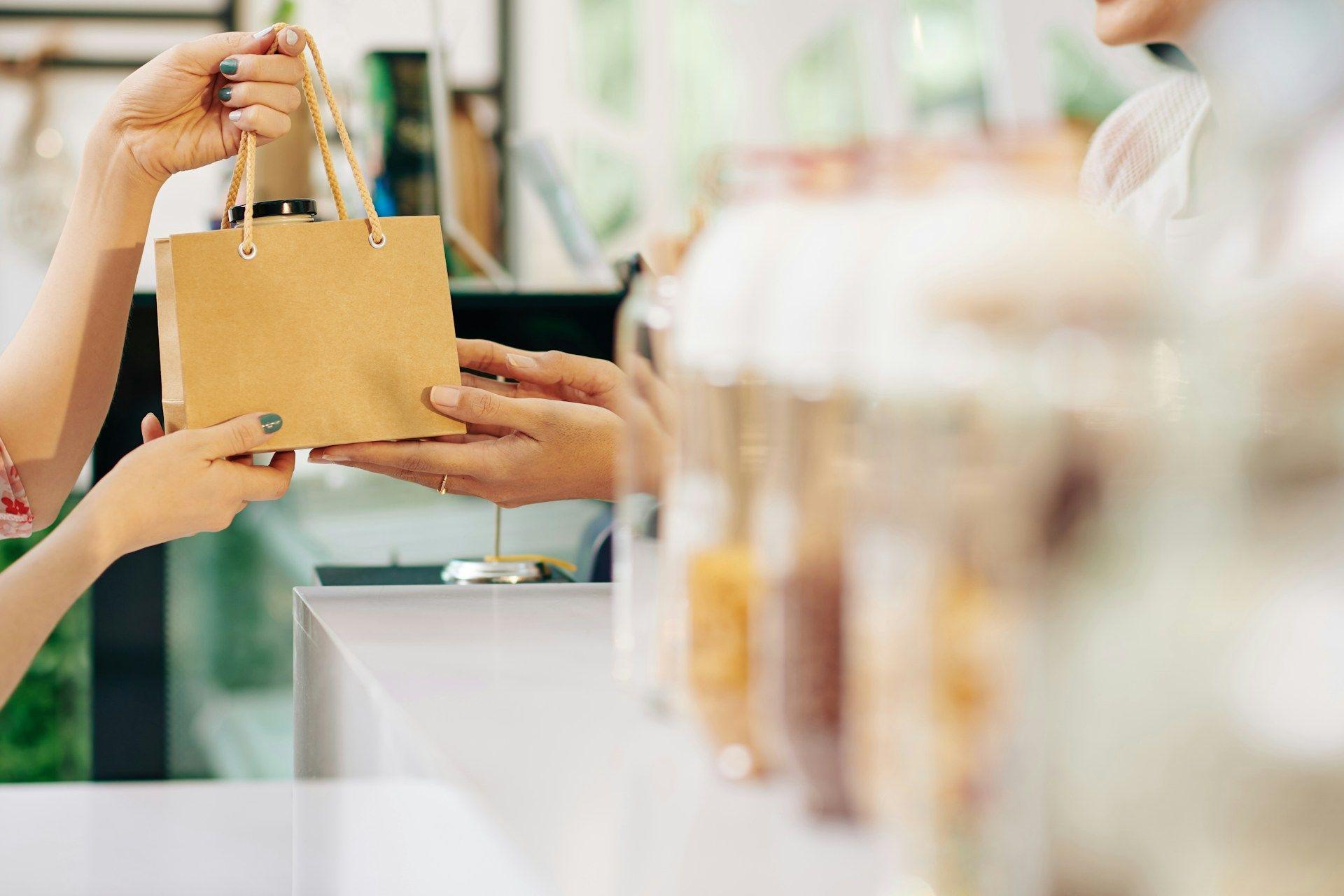 Customer's hands receiving a small brown paper bag from a shop employee.