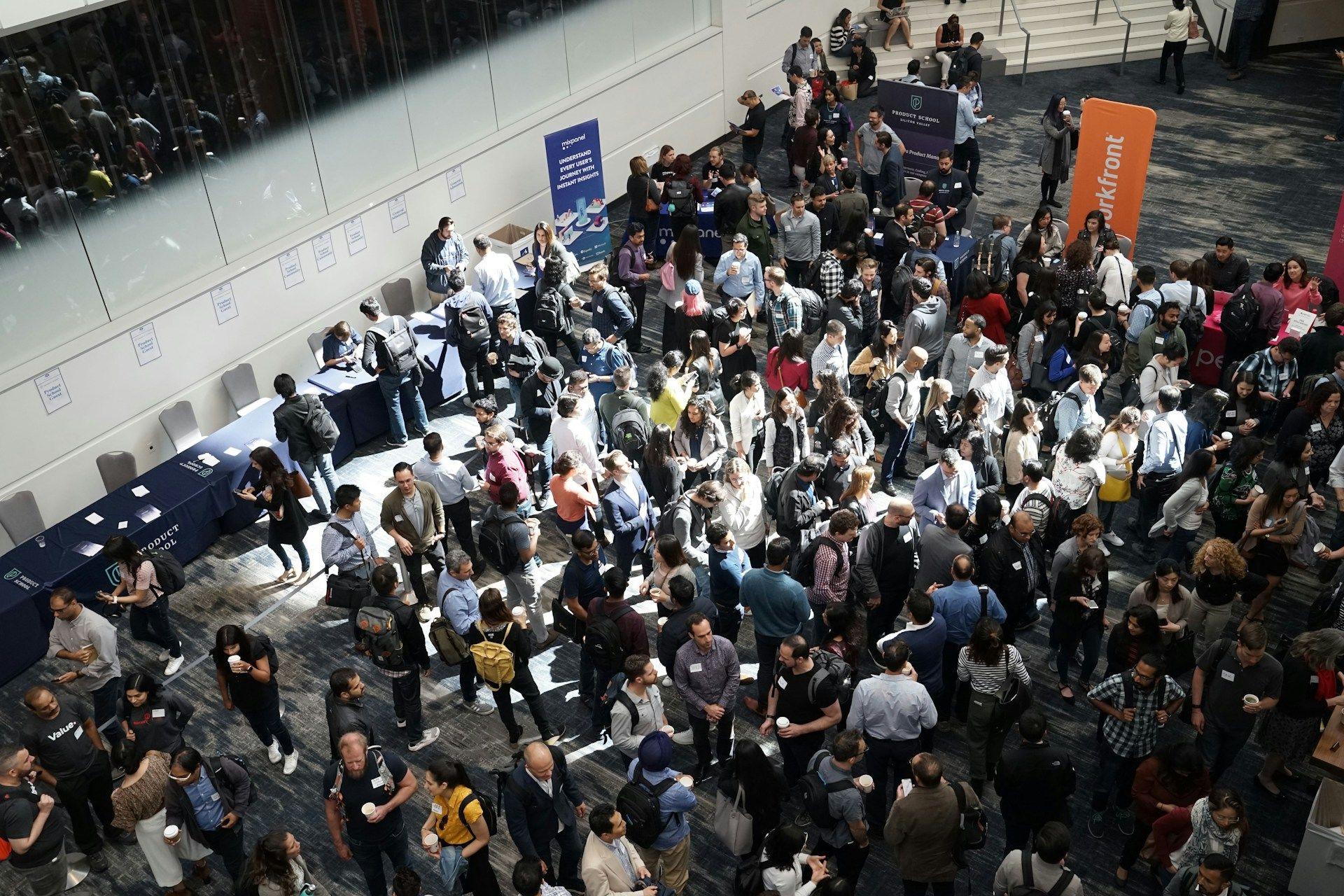 A large crowd of people mingles in a spacious indoor event hall, with some tables visible.