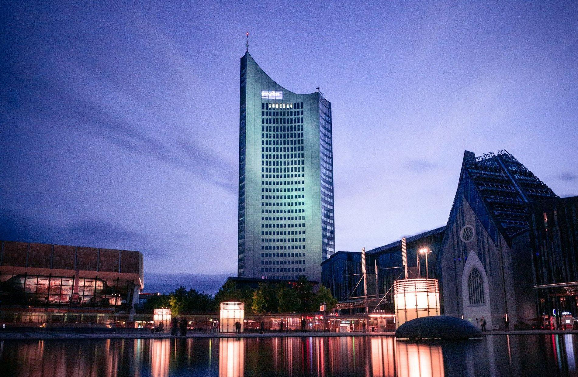 Modern skyscraper illuminated at twilight, reflected in still water with blue sky.